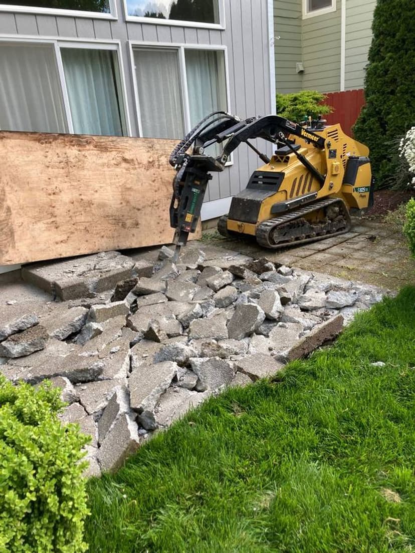 Mini excavator breaking concrete in a residential backyard with greenery nearby.